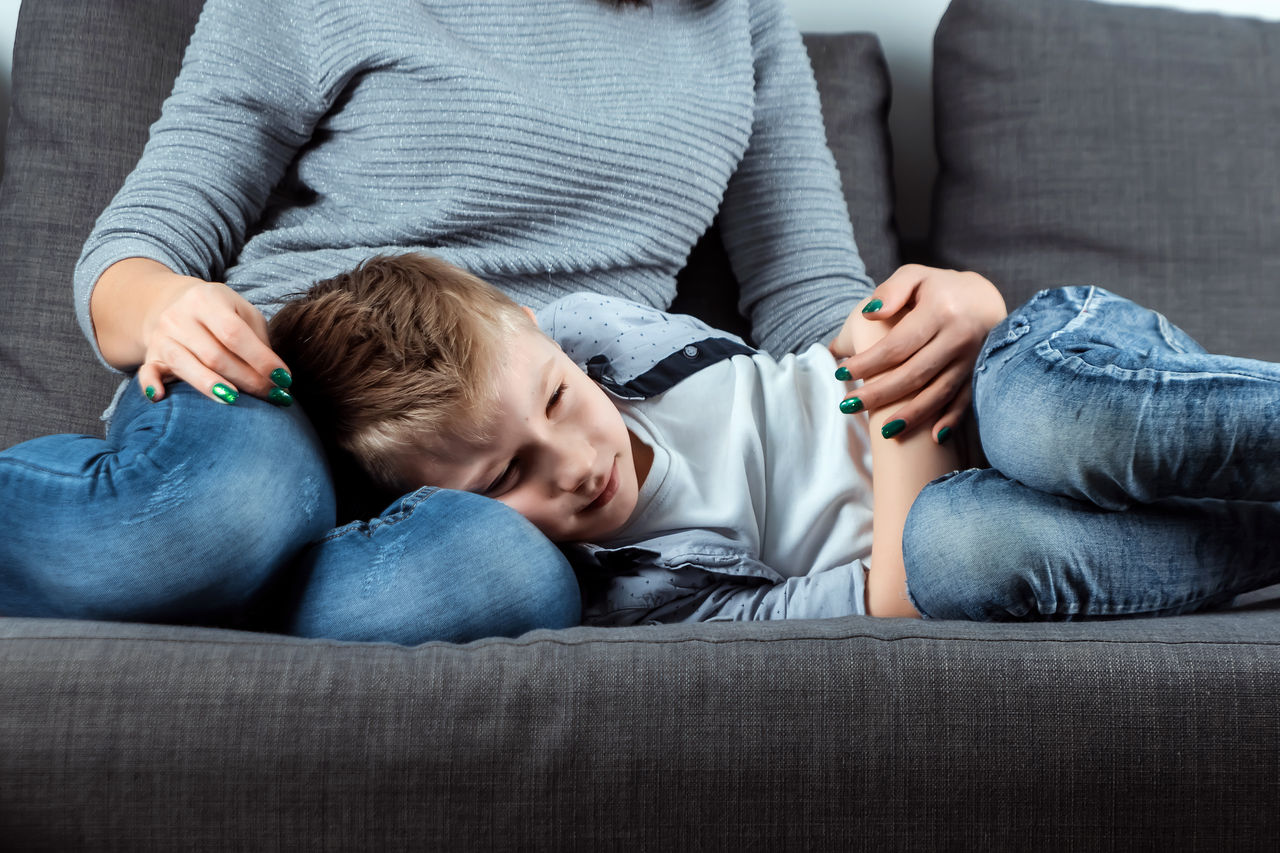 The boy is lying on the couch with an abdominal pain near his mother. The concept of custody, parental care, stomach problems, food poisoning, problems in children.