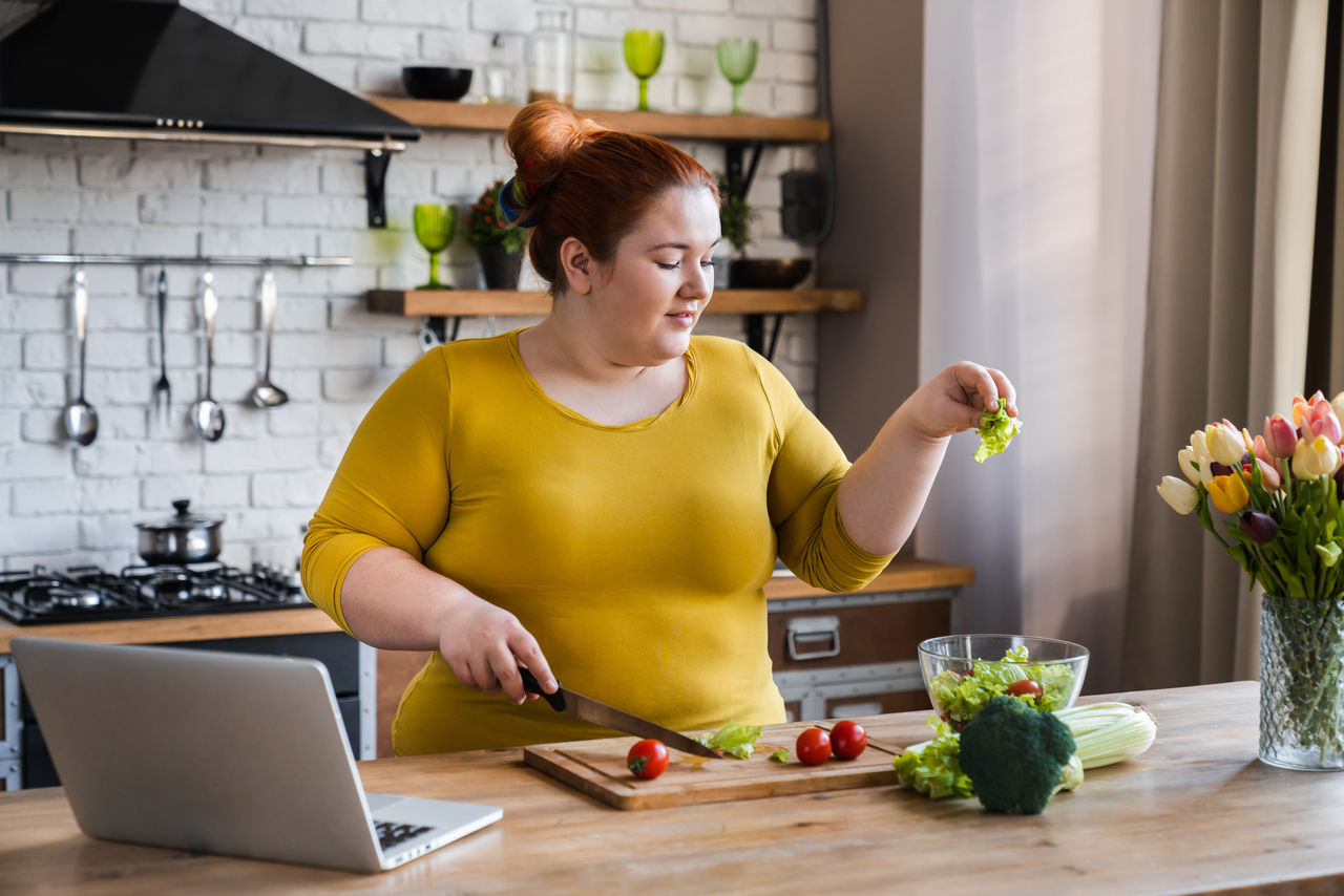 Plus size , fat caucasian woman learning to make salad and healthy food from social media adding vegetables to salad bowl ,Social distancing, stay at home concept,Plus size , fat caucasian woman learning to make salad and healt